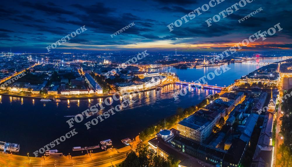 На фотографии запечатлена ночная панорама Санкт-Петербурга, освещённая огнями. Река Нева, мосты и набережные создают выразительный городской пейзаж. Видны такие достопримечательности, как Петропавловская крепость и стрелка Васильевского острова. Атмосфера белых ночей подчёркивает уникальность города.